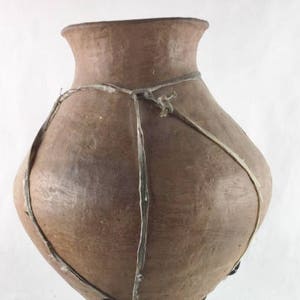 May include: A large, brown clay pot with a wide mouth and a rounded body. The pot is tied with a thick, light brown cord.