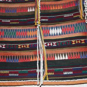 May include: A colorful woven fabric with intricate patterns in red, blue, green, yellow, and white. The fabric has a fringe of white beads and tassels.
