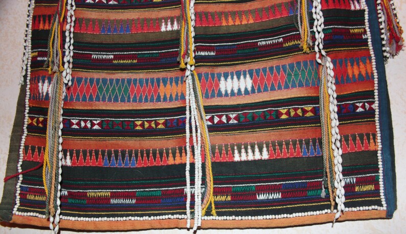 May include: A colorful woven fabric with intricate patterns in red, blue, green, yellow, and white. The fabric has a fringe of white beads and tassels.