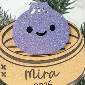 May include: A hanging ornament featuring a glittery purple dumpling character sitting inside a light-coloured wooden bamboo steamer basket. The wood is engraved with the name Mira and the year 2026, and the ornament hangs from a thin twine string.