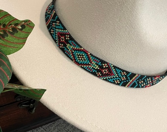 May include: A cream-colored fedora hat featuring a wide brim and a pinched crown. The hat is accented with a decorative band made of small, multi-colored beads in a geometric pattern of black, turquoise, red, and yellow, encircling the base.