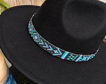 May include: A black fedora hat featuring a wide brim and a pinched crown. A decorative band made of small beads in shades of turquoise, blue, purple, and black wraps around the base of the crown, displaying a repeating geometric pattern.
