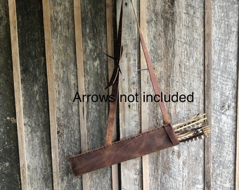May include: A brown leather quiver with a strap for carrying arrows. The quiver is hanging on a wooden wall. "Arrows not included" is written on the wall.