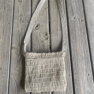 May include: A beige woven shoulder bag with a rectangular body and a long strap. The bag has a grid-like pattern and is made of a natural fiber material. The strap is also woven and attached to the top corners of the bag.
