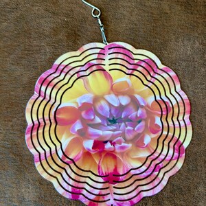 May include: A circular metal wind spinner featuring a vibrant dahlia flower print in shades of yellow, orange, and magenta. The design consists of concentric scalloped rings cut into the metal, suspended by a silver metal hook and swivel.