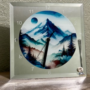 May include: Square glass clock with a mountain landscape design. The clock face features a watercolor painting of blue and teal mountains, a moon, and pine trees. The clock has black hands and silver numerals. The clock is displayed on a wooden surface.