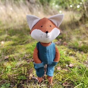 Puede incluir: Un muñeco de zorro de peluche con cara marrón y blanca, orejas beige y un mono verde azulado. El muñeco está de pie en una zona cubierta de hierba con un fondo borroso de árboles y follaje.