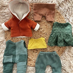 Puede incluir: Un conjunto de ropa para muñecas en varios colores. El conjunto incluye una chaqueta con capucha color óxido, pantalones cortos marrones, un vestido verde, una blusa amarilla y un peto y pantalón verdes. Las prendas están hechas de tela suave y texturizada.