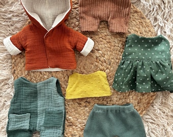 Puede incluir: Un conjunto de ropa para muñecas en varios colores. El conjunto incluye una chaqueta con capucha color óxido, pantalones cortos marrones, un vestido verde, una blusa amarilla y un peto y pantalón verdes. Las prendas están hechas de tela suave y texturizada.
