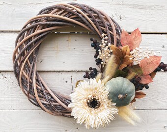May include: A woven grapevine wreath with fall-themed decorations. The wreath features a large cream-colored gerbera daisy, a small teal pumpkin, and dried leaves and berries in shades of brown, orange, and black. The wreath is set against a white wooden background.