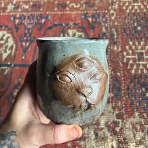 May include: A ceramic mug with a light gray, speckled glaze featuring a raised, brown, three-eyed feline face relief on the front. The mug has a rounded body and a smooth, slightly flared rim, showcasing a handcrafted, textured appearance.