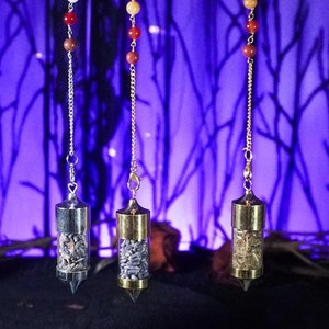May include: Three silver and glass pendulums with different colored beads hanging from a black stand against a purple background with a black silhouette of branches and birds. The pendulums are labeled "Lavender", "Lavender", and "Woodwort".