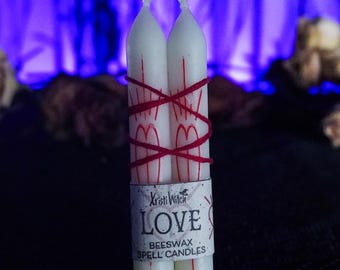 May include: Two white beeswax spell candles tied together with red string. The candles are labeled "LOVE" and "BEESWAX SPELL CANDLES" with the brand name "Kristi Witch" printed on the label.