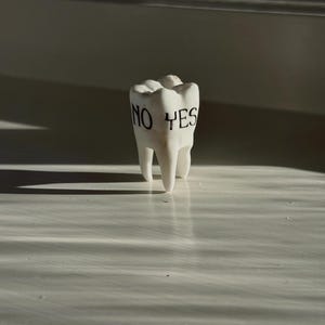 May include: A white ceramic tooth with the words "NO YES" printed in black. The tooth is standing upright on a white surface, with shadows cast across the surface.