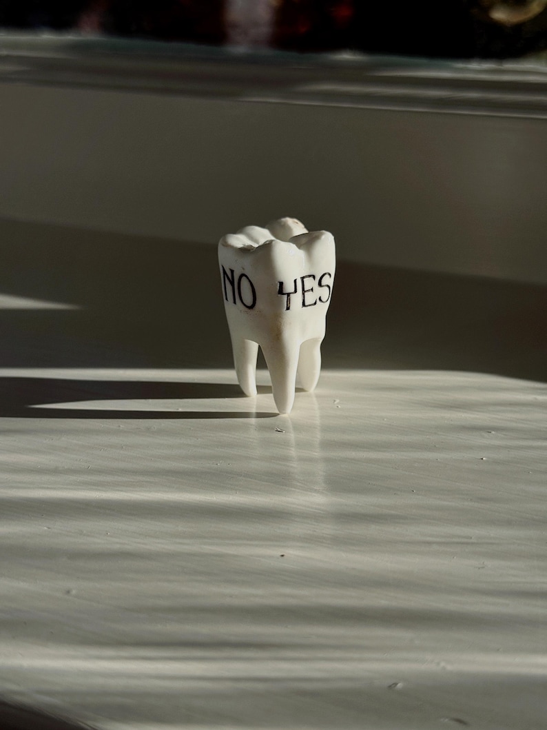 May include: A white ceramic tooth with the words "NO YES" printed in black. The tooth is standing upright on a white surface, with shadows cast across the surface.