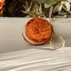 Puede incluir: Un objeto redondo, de color marrón anaranjado, con una superficie texturizada, posiblemente un elemento decorativo o natural. Está atado con hilo natural, lo que sugiere una estética rústica o artesanal. El objeto se encuentra sobre una superficie blanca.