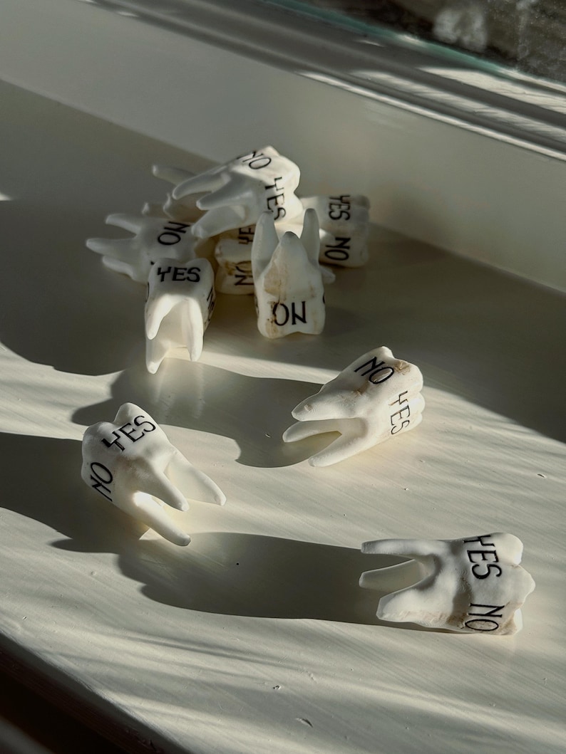 May include: A collection of white ceramic teeth, each inscribed with the words "YES" and "NO" in black lettering. The teeth are scattered on a white surface, with shadows cast by sunlight.