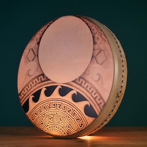 May include: A round, wooden frame drum with a leather head. The head is decorated with a sun and moon design in black, white, and brown. The design includes a stylized sun with rays and a crescent moon with a geometric pattern.