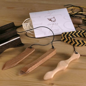 Puede incluir: Un conjunto de herramientas de madera para instrumentos de cuerda, que incluye un enrollador de cuerdas, un extractor de clavijas de puente y una cejilla. Las herramientas se muestran sobre una superficie de madera con una correa trenzada negra y dorada y un paquete de cuerdas.