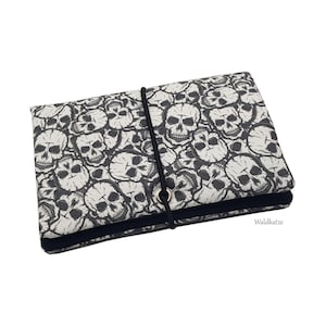 Puede incluir: Un estuche rectangular de tela con un patrón repetitivo de calaveras en blanco y negro. El estuche se asegura con un cordón elástico negro que pasa por un ojal metálico plateado. El texto Waldkatze aparece en la esquina inferior derecha.