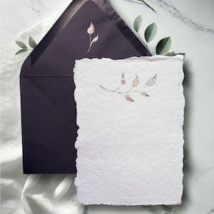 May include: A stationery set featuring a textured white paper with a deckled edge, a matching dark purple envelope, and a sprig design with metallic accents. The set is ideal for invitations or personalized notes.