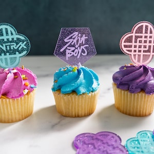 May include: Three cupcakes with colorful frosting and decorative toppers. The cupcakes have pink, blue, and purple frosting, with sprinkles. The toppers are shaped with the text "HUNTRAX" and "SAJA BOYS".