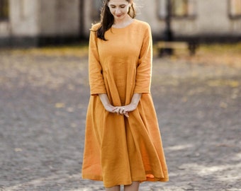 May include: A woman in a mustard yellow dress stands on a cobblestone street. She is looking down at her feet and her hands are clasped in front of her. The background is a blurry image of a building with trees.