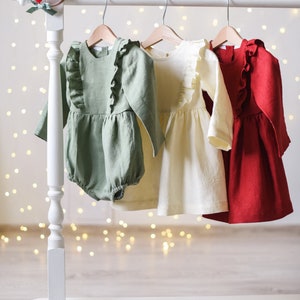 May include: Three baby dresses hanging on a white rack. The dresses are made of linen and have ruffles. The dresses are green, cream, and red.
