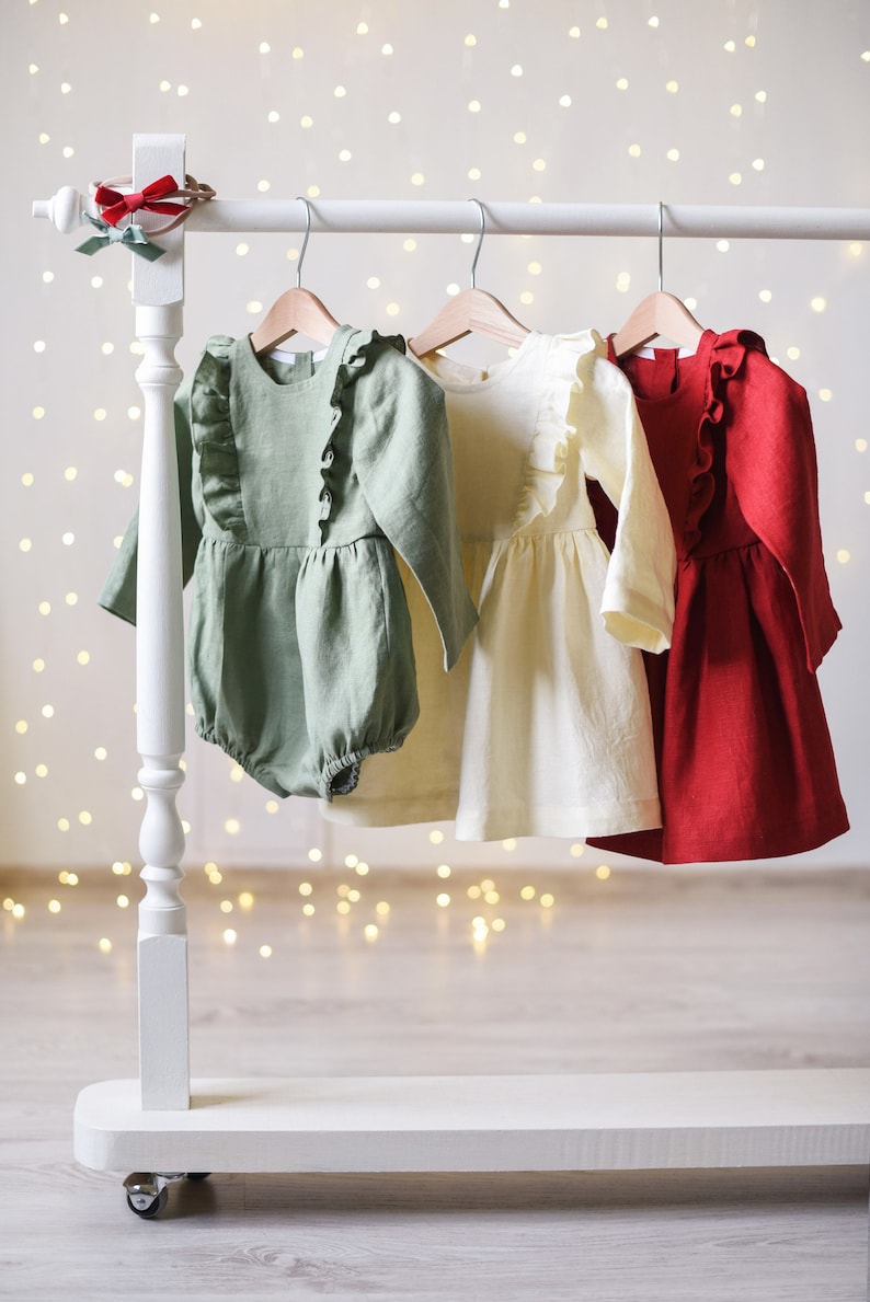 May include: Three baby dresses hanging on a white rack. The dresses are made of linen and have ruffles. The dresses are green, cream, and red.