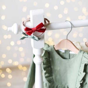 May include: A white wooden clothing rack with a green dress hanging on a wooden hanger. The dress has ruffles around the neckline and sleeves. There are two hair bows on the rack, one red and one green.