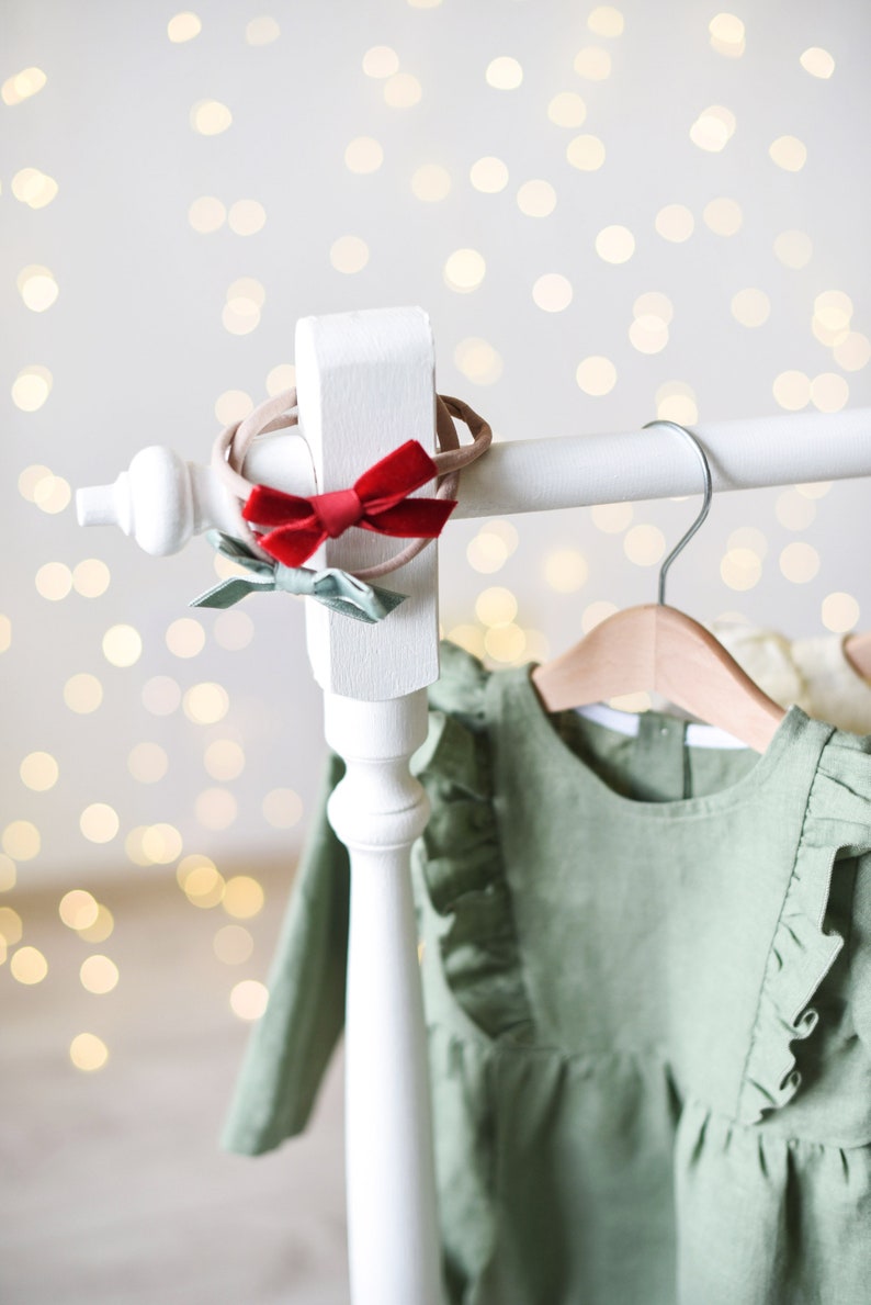 May include: A white wooden clothing rack with a green dress hanging on a wooden hanger. The dress has ruffles around the neckline and sleeves. There are two hair bows on the rack, one red and one green.