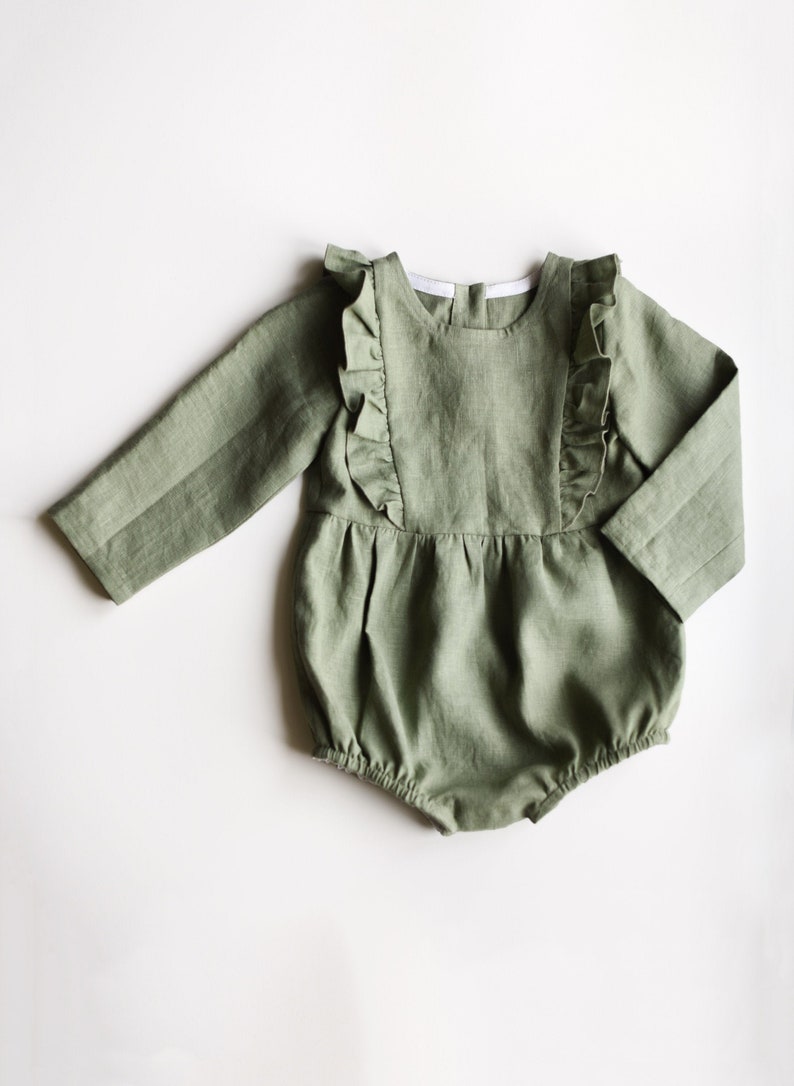 May include: A sage green linen baby romper with ruffled detailing around the neckline and shoulders. The romper has long sleeves and a gathered skirt.