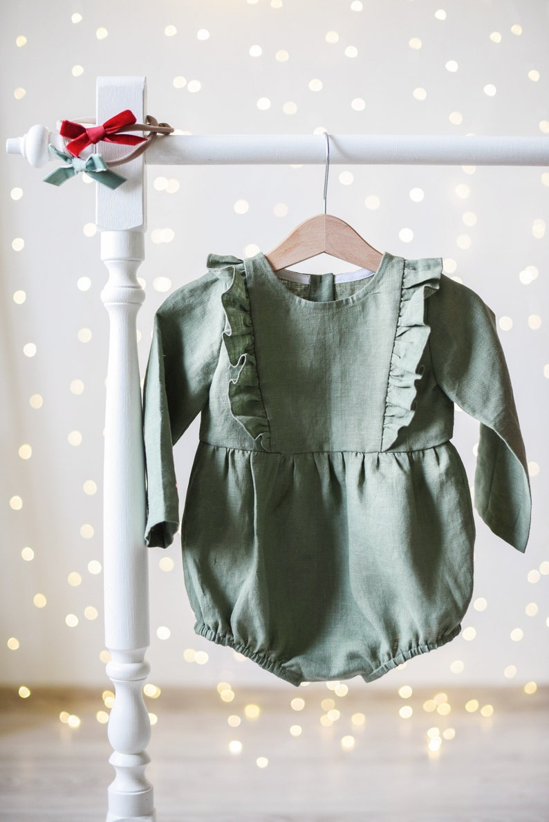 May include: A sage green linen baby romper with ruffled detailing on the shoulders and sleeves. The romper is hanging on a white wooden hanger against a white background with bokeh lights.