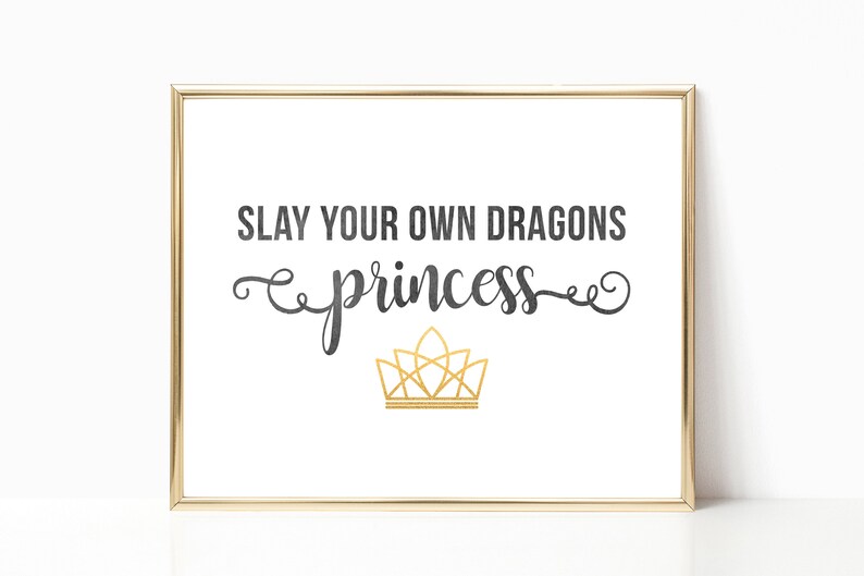 May include: A gold framed print with the text "SLAY YOUR OWN DRAGONS princess" in black with a gold crown underneath.
