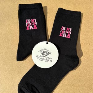 May include: Black socks with the text "IN MY FORTIES ERA" in pink. A white tag with the text "Louise Martin Julie Personalised Diamante" is placed between the socks.
