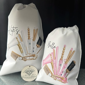 Pode incluir: Duas bolsas de lona brancas com nomes personalizados, "Kate's" e "Bella", que apresentam ilustra&ccedil;&otilde;es de ferramentas para pentear o cabelo, incluindo um secador de cabelo, um modelador de cachos, uma prancha e escovas. As bolsas s&atilde;o decoradas com detalhes dourados e rosados.