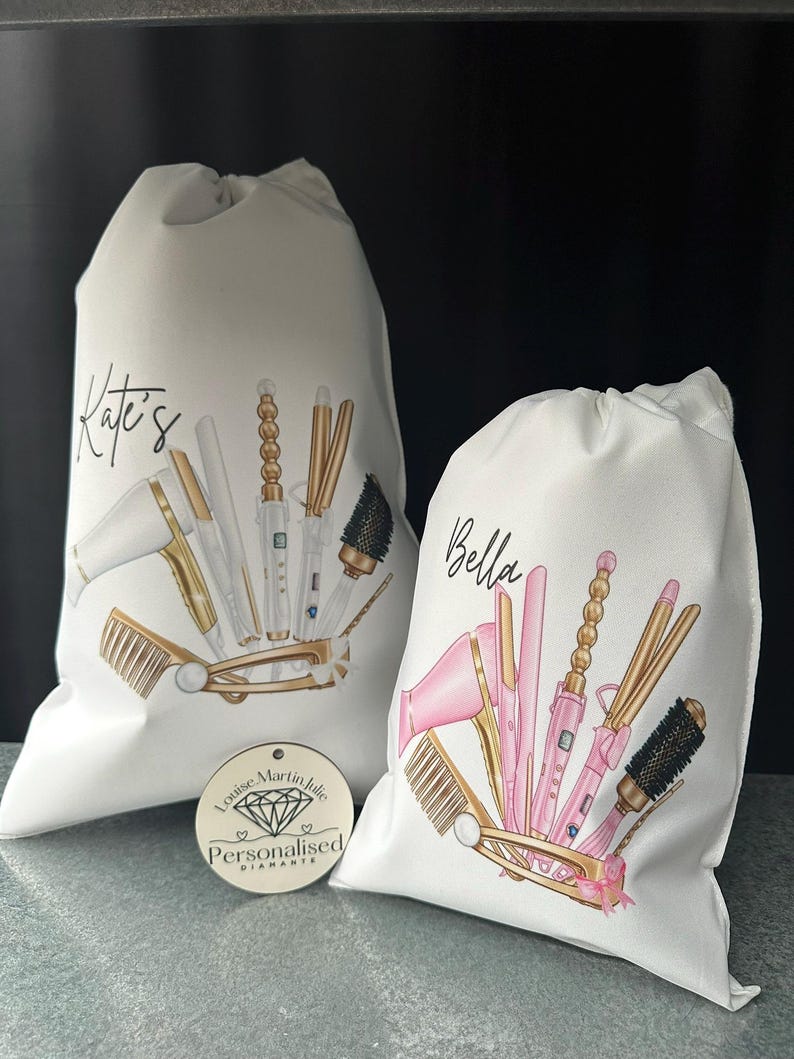 Pode incluir: Duas bolsas de lona brancas com nomes personalizados, "Kate's" e "Bella", que apresentam ilustra&ccedil;&otilde;es de ferramentas para pentear o cabelo, incluindo um secador de cabelo, um modelador de cachos, uma prancha e escovas. As bolsas s&atilde;o decoradas com detalhes dourados e rosados.