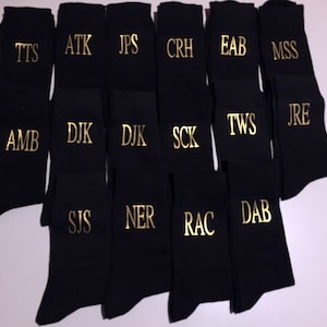 May include: A collection of black socks with gold embroidered initials. The initials include TTS, ATK, JPS, CRH, EAB, MSS, AMB, DJK, SCK, TWS, JRE, SIS, NER, RAC, and DAB.