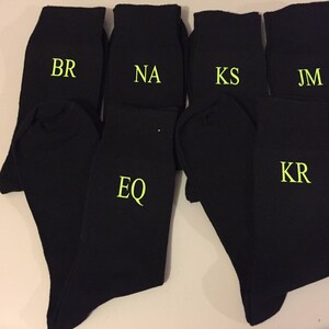 May include: Six black socks with different initials embroidered in yellow on the front. The initials are BR, NA, KS, JM, EQ, and KR.