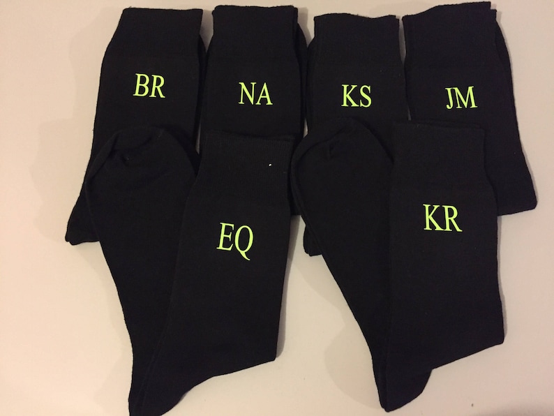 May include: Six black socks with different initials embroidered in yellow on the front. The initials are BR, NA, KS, JM, EQ, and KR.