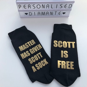 Pode incluir: Um par de meias pretas com letras douradas. Uma meia diz "Master has given Scott a sock" e a outra diz "Scott is free".