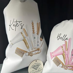 Pode incluir: Duas bolsas de lona brancas com ferramentas de penteado douradas e cor-de-rosa impressas nelas. As bolsas s&atilde;o personalizadas com os nomes "Kate's" e "Bella". Uma etiqueta redonda de madeira com o texto "Louise Martin Julie Personalizado Diamante" est&aacute; no canto inferior direito da imagem.