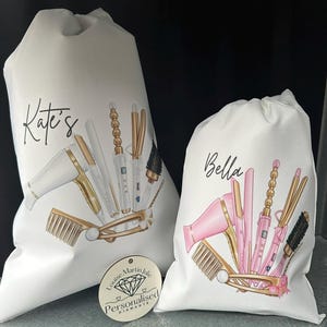 Pode incluir: Duas bolsas brancas de cord&atilde;o com os nomes personalizados "Kate's" e "Bella" impressos nelas. As bolsas apresentam ilustra&ccedil;&otilde;es de ferramentas para pentear o cabelo, como um secador de cabelo, um modelador de cachos e um pente. Uma pequena etiqueta de madeira com o texto "Louise Martin Julie Personalised Diamante" &eacute; vis&iacute;vel no canto inferior esquerdo.