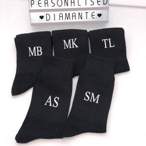May include: Five pairs of black socks with white embroidered initials: MB, MK, TL, AS, and SM. The socks are arranged in a pile on a white surface. The text "PERSONALISED DIAMANTE" is visible in the background.