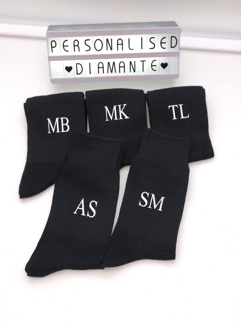 May include: Five pairs of black socks with white embroidered initials: MB, MK, TL, AS, and SM. The socks are arranged in a pile on a white surface. The text "PERSONALISED DIAMANTE" is visible in the background.