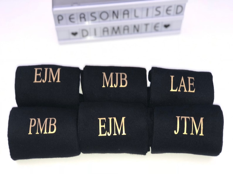 May include: Six pairs of black socks with gold embroidered initials. The initials are EJM, MJB, LAE, PMB, EJM, and JTM.