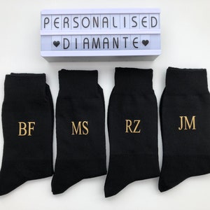 May include: Four black socks with gold embroidered initials: BF, MS, RZ, and JM. The socks are arranged in a row on a white surface. A white light box with the text "Personalised Diamante" is above the socks.