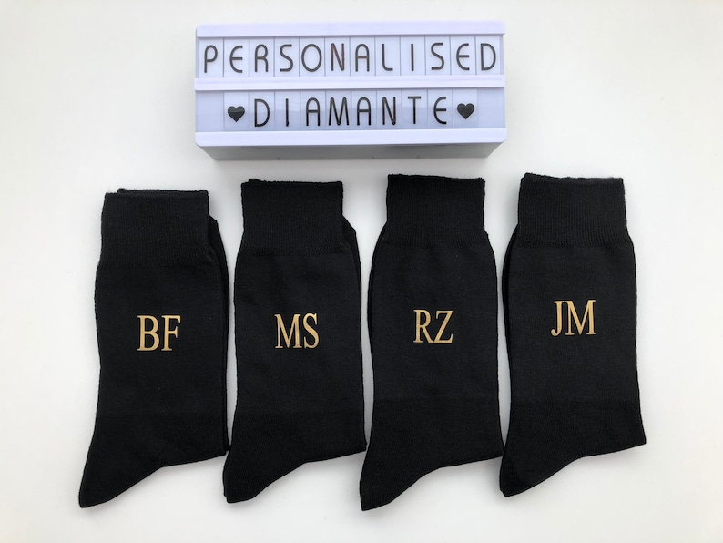 May include: Four black socks with gold embroidered initials: BF, MS, RZ, and JM. The socks are arranged in a row on a white surface. A white light box with the text "Personalised Diamante" is above the socks.