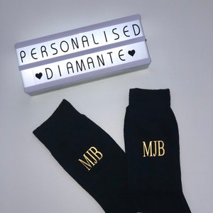 May include: A pair of black socks with gold embroidered initials "MJB". The socks are displayed next to a white light-up sign with the text "PERSONALISED &hearts; DIAMANTE".