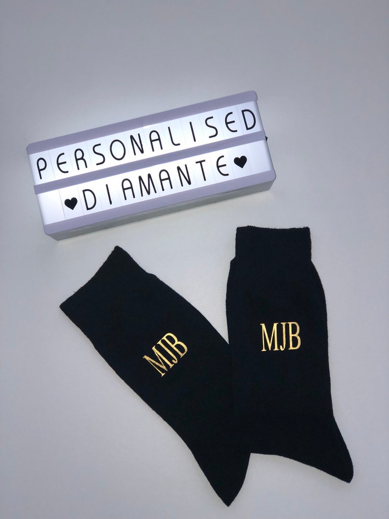 May include: A pair of black socks with gold embroidered initials "MJB". The socks are displayed next to a white light-up sign with the text "PERSONALISED &hearts; DIAMANTE".